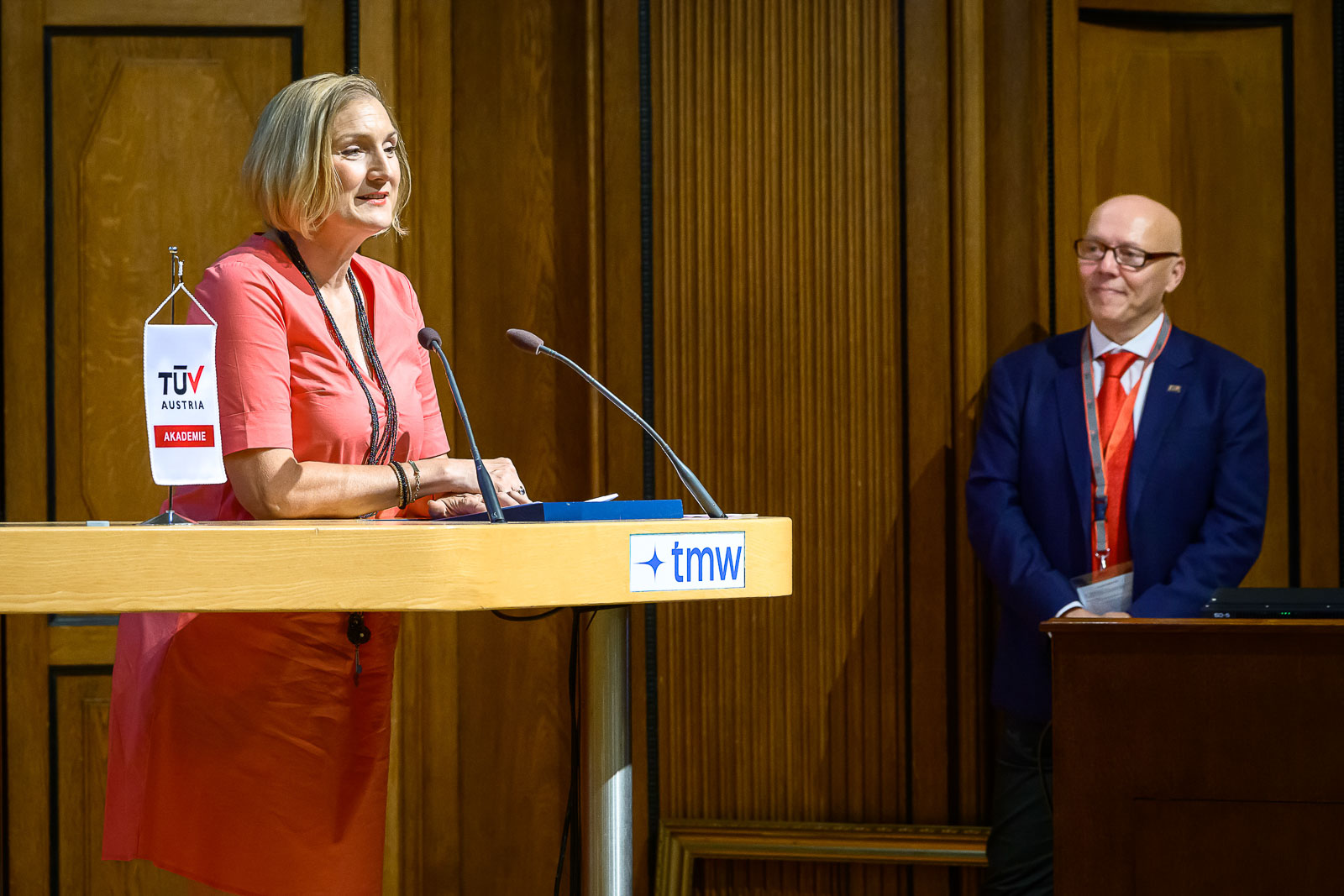 <br /> Dame steht am Rednerpult und spricht. Ein Mann im Anzug steht daneben.